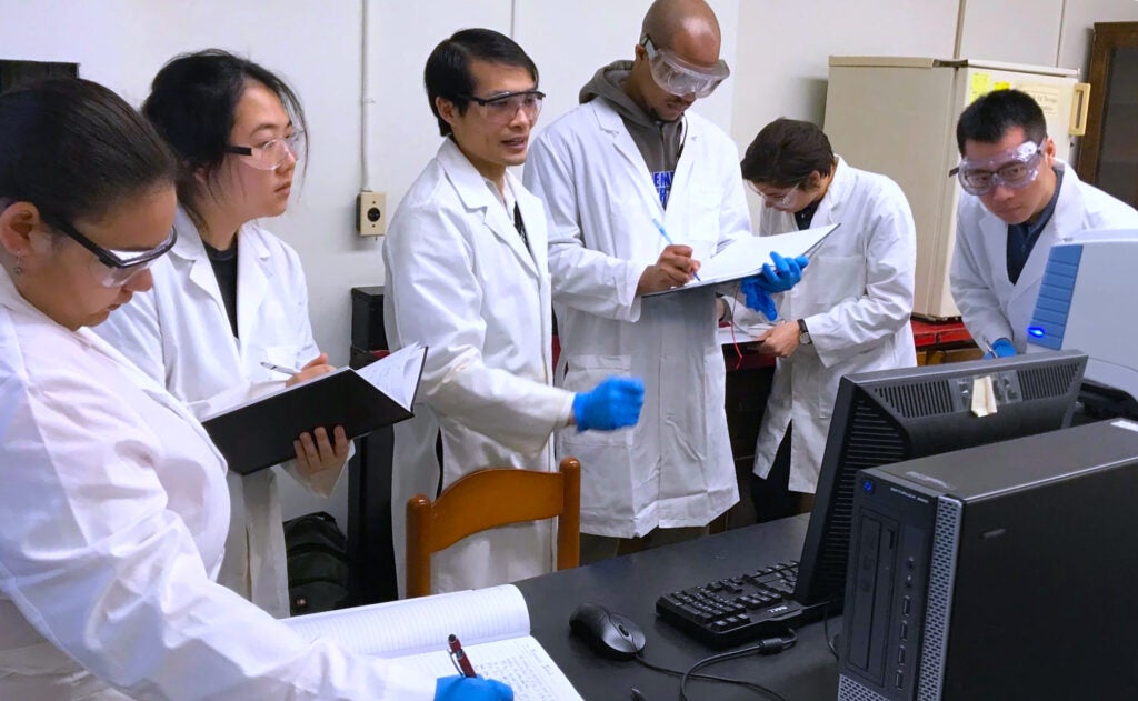 students discussing and taking notes in lab coats at a computer