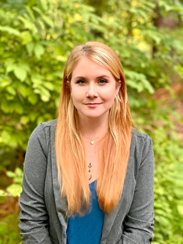 Hanna Hidle wearing a blue shirt and a grey sweater against a leafy background